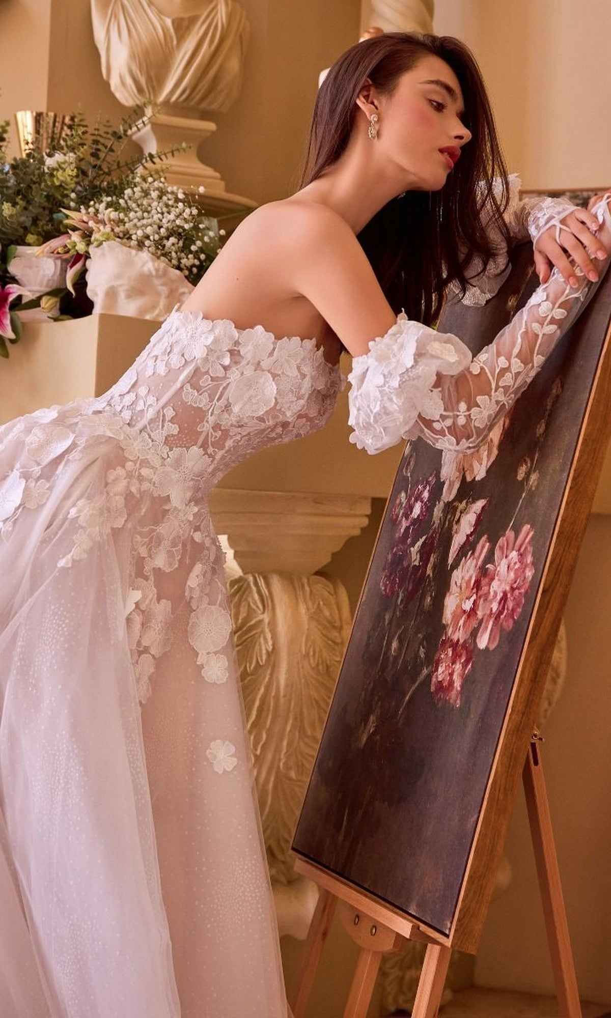A woman in the Andrea and Leo WL017 long wedding dress, featuring 3D floral appliques, leans on an easel with a pink and red flower painting, surrounded by classical decor.